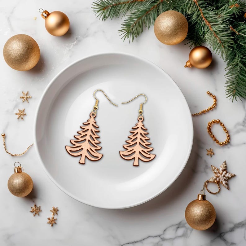 May include: Two wooden Christmas tree earrings with gold-colored hooks rest on a white plate. The earrings are surrounded by gold ornaments, snowflakes, and a sprig of evergreen. The earrings are a festive holiday accessory.