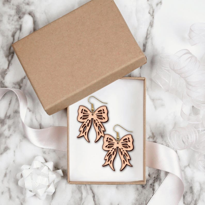 May include: A pair of light brown wooden bow-shaped earrings with gold-colored hooks, presented in an open brown gift box. The earrings have a detailed, openwork design. The box sits on a marble surface, with a white ribbon and bow.