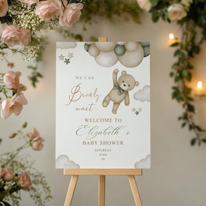 May include: A baby shower welcome sign on an easel. The sign features a teddy bear illustration with the text "We can bearly wait" and "Welcome to Elizabeth's Baby Shower". The sign is decorated with clouds and stars.