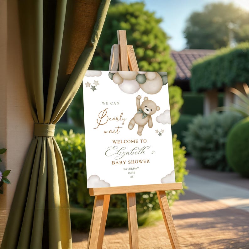 May include: A baby shower welcome sign on a wooden easel. The sign features a teddy bear illustration with the text "We can bearly wait" and "Welcome to Elizabeth's Baby Shower" with the date Saturday, June 28.