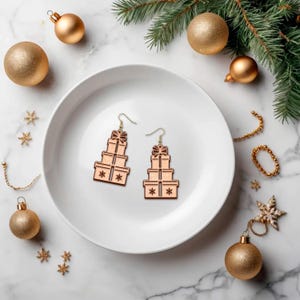 May include: Wooden Christmas gift box earrings with a bow and snowflake design. The earrings are light brown and are displayed on a white plate with gold Christmas ornaments and a sprig of evergreen. Festive holiday jewelry.