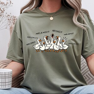 May include: Olive green t-shirt featuring a cartoon illustration of several white geese with orange beaks and feet. The geese are yelling, with the text "MRS.EMILY!" above them. The shirt has a crew neck.