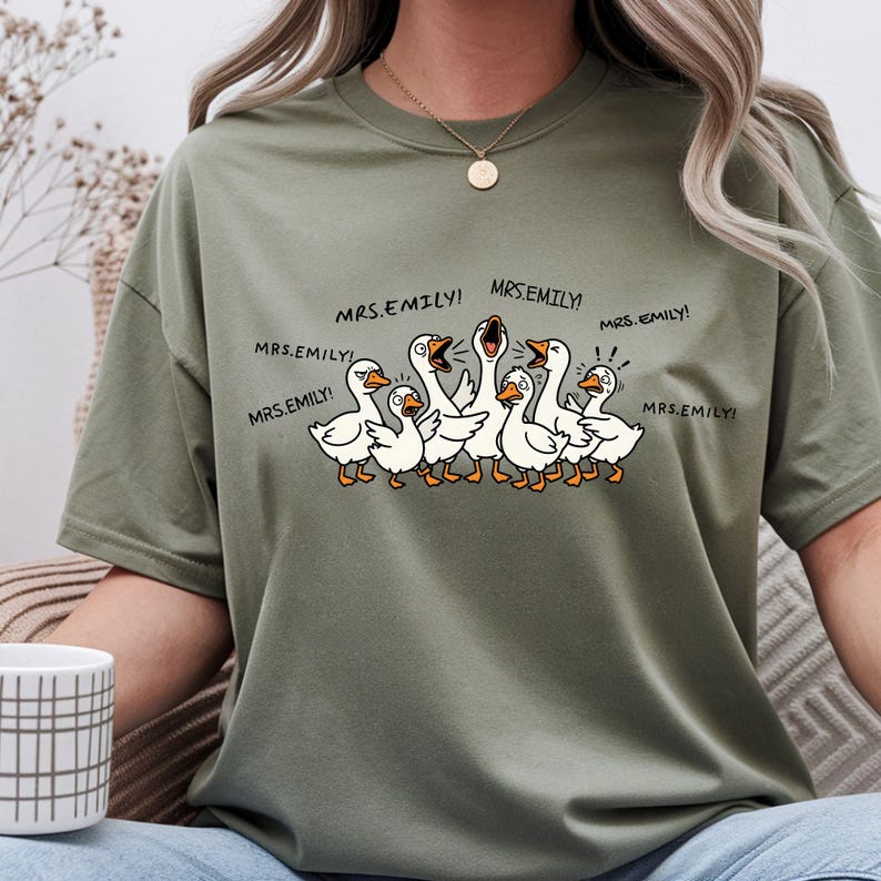 May include: Olive green t-shirt featuring a cartoon illustration of several white geese with orange beaks and feet. The geese are yelling, with the text "MRS.EMILY!" above them. The shirt has a crew neck.