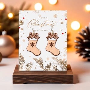 May include: Wooden Christmas stocking earrings with a bow and snowflake design, displayed on a greeting card that says "Merry Christmas". The earrings are light brown and hang from gold-colored hooks. The card is on a dark wooden stand.