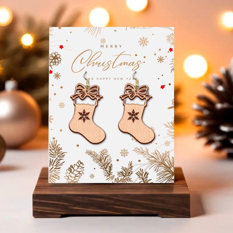 May include: Wooden Christmas stocking earrings with a bow and snowflake design, displayed on a greeting card that says "Merry Christmas". The earrings are light brown and hang from gold-colored hooks. The card is on a dark wooden stand.