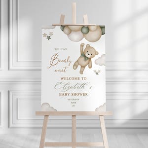 May include: A baby shower welcome sign on a wooden easel. The sign features a teddy bear illustration with the text "We can bearly wait" and "Welcome to Elizabeth's Baby Shower". The date is June 28.