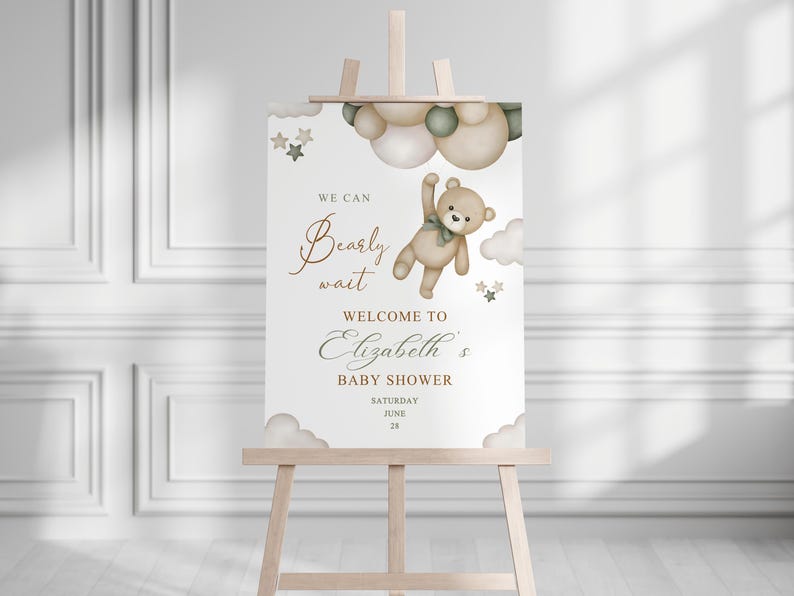 May include: A baby shower welcome sign on a wooden easel. The sign features a teddy bear illustration with the text "We can bearly wait" and "Welcome to Elizabeth's Baby Shower". The date is June 28.