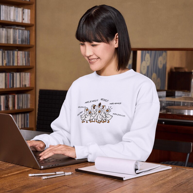 May include: White sweatshirt featuring a cartoon illustration of geese with speech bubbles. The geese are drawn in a simple style with brown outlines and details. Text includes "Mrs. Emily!" and other names.