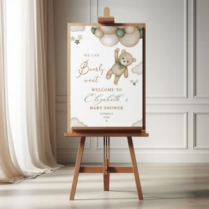May include: A wooden easel displays a baby shower welcome sign. The sign features a teddy bear illustration with the text "We can bearly wait" and "Welcome to Elizabeth's Baby Shower". The sign is decorated with clouds and stars.
