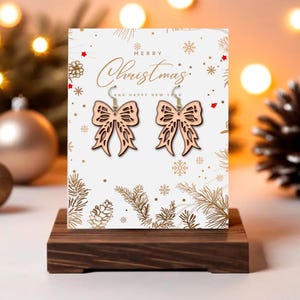 May include: Wooden bow-shaped earrings with gold-colored hooks are displayed on a Christmas-themed card. The card reads "Merry Christmas and Happy New Year" with festive illustrations. The earrings are on a dark wooden stand.