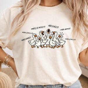 May include: Cream-colored t-shirt featuring a cartoon illustration of a flock of white geese with orange beaks and feet. The geese are yelling, with the text "MRS.EMILY!" repeated around them. The shirt has a relaxed fit.