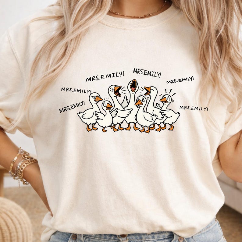 May include: Cream-colored t-shirt featuring a cartoon illustration of a flock of white geese with orange beaks and feet. The geese are yelling, with the text "MRS.EMILY!" repeated around them. The shirt has a relaxed fit.