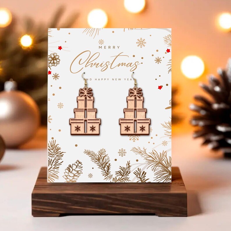 May include: Wooden Christmas earrings shaped like stacked gift boxes with bow details. The earrings are displayed on a white card with the words "Merry Christmas" and festive decorations. The earrings are on a wooden display stand.