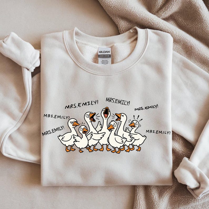 May include: A cream-colored sweatshirt featuring a cartoon illustration of a flock of white geese with orange beaks and feet. The geese are yelling, with the text "MRS. EMILY!" repeated around them. A Gildan tag is visible.