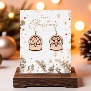 May include: Wooden snowflake earrings displayed on a Christmas-themed card. The earrings are light brown, shaped like snow globes with snowflake cutouts. The card reads "Merry Christmas and Happy New Year" with gold accents and snowflake illustrations. The earrings are on a wooden stand.