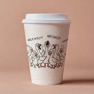 May include: A white paper coffee cup with a white lid. The cup features a cartoon illustration of a flock of geese with open beaks and the text "MRS.EMILY!". The background is a solid light brown.
