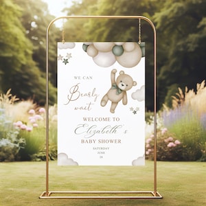 May include: A baby shower welcome sign in a gold frame. The sign features a teddy bear illustration with the text "We can bearly wait" and "Welcome to Elizabeth's Baby Shower". The background includes clouds and stars.