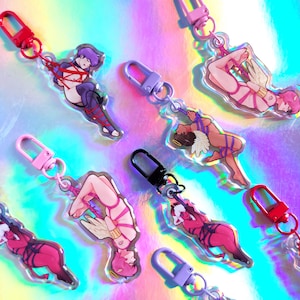 Op de afbeelding: Zes kleurrijke sleutelhangers met verschillende personages die vastgebonden zijn met touw. Elke sleutelhanger heeft een metalen sluiting en een transparante achtergrond. De personages zijn een vrouw met paars haar, een vrouw met roze haar en engelenvleugels, een man met engelenvleugels, een vrouw met engelenvleugels, een vrouw met duivelshoorns en een man met duivelshoorns.