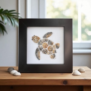 Puede incluir: Obra de arte enmarcada con un diseño de tortuga marina. La tortuga está hecha de pequeñas piedras texturizadas en tonos marrón, gris y beige, sobre un fondo blanco. La obra está en un marco negro y descansa sobre una superficie de madera.