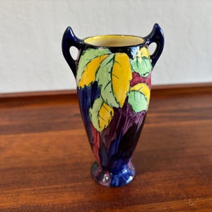 May include: A small ceramic vase featuring two dark blue handles and a tapered body. The exterior is decorated with a hand-painted pattern of bright yellow and light green leaves set against a deep purple and dark blue background with a cream interior.