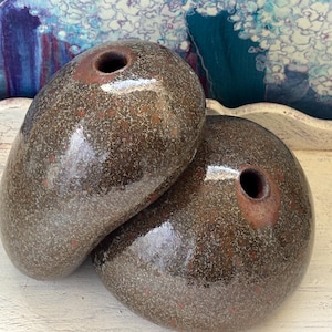 May include: Two rounded, pebble-shaped ceramic vases with a glossy, speckled brown and dark gray glaze. Each vase features a small, circular opening at the top with a reddish-brown rim, showcasing a smooth, organic form and a reflective surface.