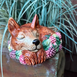 May include: A ceramic fox figurine in a sleeping pose, with closed eyes and a black nose. The fox is brown with white and tan accents. It is adorned with a decorative collar of pink, green, and purple berries. The figurine is placed in front of green foliage.