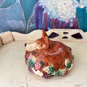 May include: A ceramic fox figurine with a reddish-brown coat, adorned with a wreath of green leaves and pink and purple berries. The fox is lying down, with its head resting on its paws. The figurine is on a white tray.