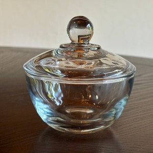May include: A clear glass candy dish with a lid. The dish has a rounded bowl shape and a lid with a small, round knob. The glass is transparent, allowing light to pass through. The dish is likely used for storing small items.