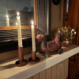 May include: Three white candles with wooden candle holders on a white mantel. A wooden horse figurine with red paint is in front of the candles.