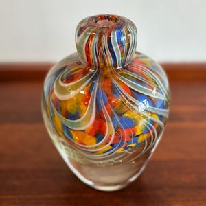 May include: A colorful glass vase with a rounded body and a narrow neck. The vase features a swirl design with shades of red, yellow, blue, and white. The vase is sitting on a wooden surface.