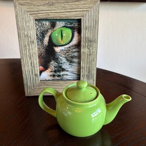 May include: A green ceramic teapot with a matching lid sits on a dark wooden surface. Behind it, a framed picture displays a close-up of a cat's eye. The eye is a vibrant green, contrasting with the gray and brown fur.