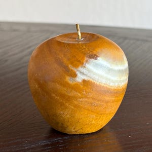 May include: A polished stone apple sculpture featuring swirling patterns of amber, brown, and white. The smooth, rounded object has a small, thin, metallic stem protruding from the center of the top indentation, showcasing natural mineral banding.