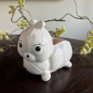 May include: A white ceramic planter in the shape of a cartoon animal, possibly a squirrel or bunny. The planter has large black eyes, a small nose, and a textured body. The planter is on a dark surface with yellow and green floral accents.