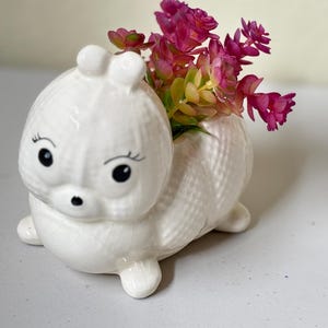 May include: A white ceramic animal-shaped planter with a textured surface. The planter has black eyes, a small black nose, and pink flowers. The planter is a decorative item for home decor.