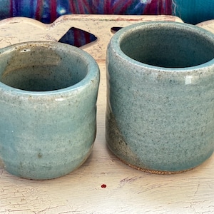 May include: Two handmade ceramic cups in a speckled, light blue glaze. The cup on the left is shorter and wider, while the one on the right is taller and more cylindrical. Both have a rustic, organic feel.