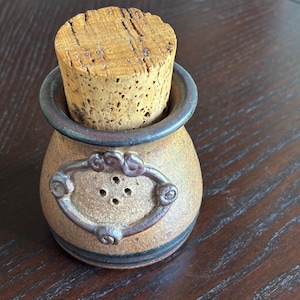 May include: A small, tan ceramic jar with a cork stopper. The jar has a decorative metal frame with three small holes and a dark brown rim. The jar is sitting on a dark brown wooden surface.