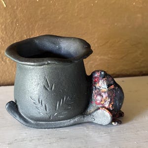 May include: A small, matte charcoal-gray ceramic vessel with a wavy, uneven rim. The exterior features a subtle, etched leaf pattern. Attached to the side is a sculpted, dark-colored creature with mottled red and orange accents on its back.
