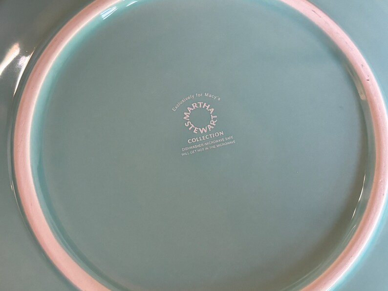 May include: A light blue ceramic plate with a white rim. The bottom of the plate has a white logo that reads "Exclusively for Macy's Martha Stewart Collection Dishwasher/Microwave Safe Will Get Hot in the Microwave".