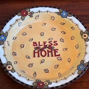 May include: A decorative ceramic plate with a cream-colored center featuring the words "BLESS HOME" in burgundy. The plate is adorned with floral and leaf motifs in red, blue, and yellow, and has a scalloped edge.