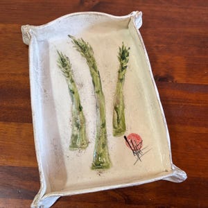 May include: A rectangular ceramic tray with a white, textured glaze and raised, pinched corners. The center features a hand-painted illustration of three green asparagus stalks and a small red circular mark with black ink-like lines.