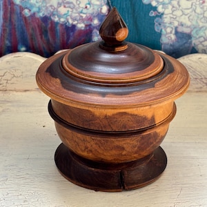 May include: A handcrafted wooden jar with a lid. The jar is made of dark and light brown wood, with a rounded base and a conical finial on the lid. The wood grain is visible, adding to its rustic charm. The jar is likely used for storage.