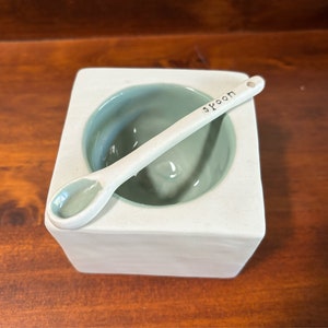 May include: A square ceramic container with a matte white exterior and a smooth, light sage green interior. A matching ceramic spoon rests diagonally across the top, featuring the word "spoon" stamped in black lettering on its handle.