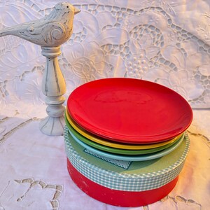May include: A set of four colorful ceramic dessert plates stacked in a box. The plates are red, yellow, green, and blue. The box is white with a blue and white checkered pattern.