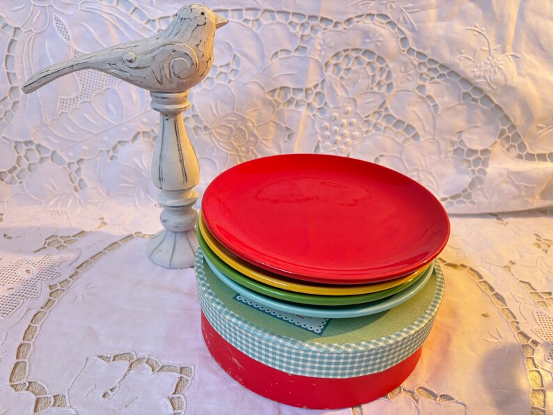 May include: A set of four colorful ceramic dessert plates stacked in a box. The plates are red, yellow, green, and blue. The box is white with a blue and white checkered pattern.