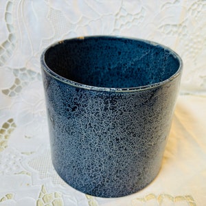 May include: A cylindrical ceramic container with a speckled navy blue glaze. The container has a slightly flared rim and a textured surface. The background is a white lace tablecloth.