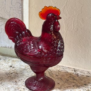 May include: A ruby red glass rooster-shaped covered dish featuring a textured feather pattern on the body and a vibrant orange-red comb. The rooster stands on a circular pedestal base and includes a removable lid that forms the upper body.