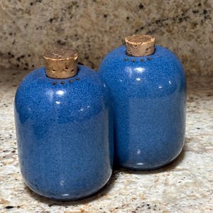May include: Two blue ceramic salt and pepper shakers with cork stoppers. The shakers have a speckled finish and a rounded shape. The cork stoppers are light brown and cylindrical.