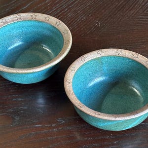 May include: Two handmade ceramic bowls with a turquoise interior and a tan rim. The rim is decorated with small star shapes. The bowls are sitting on a dark wooden surface.