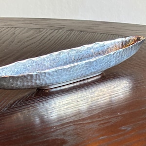 May include: A long, oval-shaped decorative bowl with a hammered texture. The bowl has a light blue and silver finish with a bronze-colored rim. The bowl is resting on a dark wooden surface.