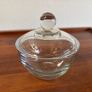 May include: A clear glass candy dish with a lid. The dish is round with a small knob on top of the lid. The dish is sitting on a wooden surface.